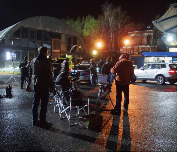 A nighttime film set scene with crew members, filming equipment, lights, and vehicles outside industrial buildings.