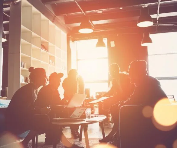 A group of people is gathered around a table in a modern office space, discussing work with laptops and documents.