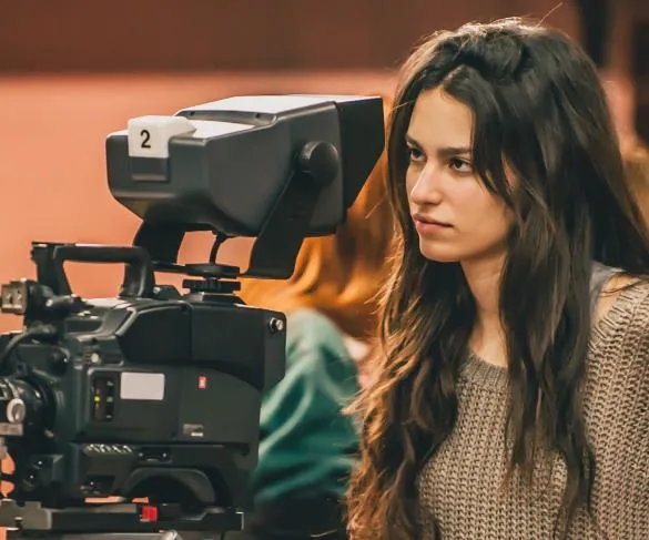 A woman stands behind and looks into a professional camera, wearing a brown sweater, with a background out of focus.