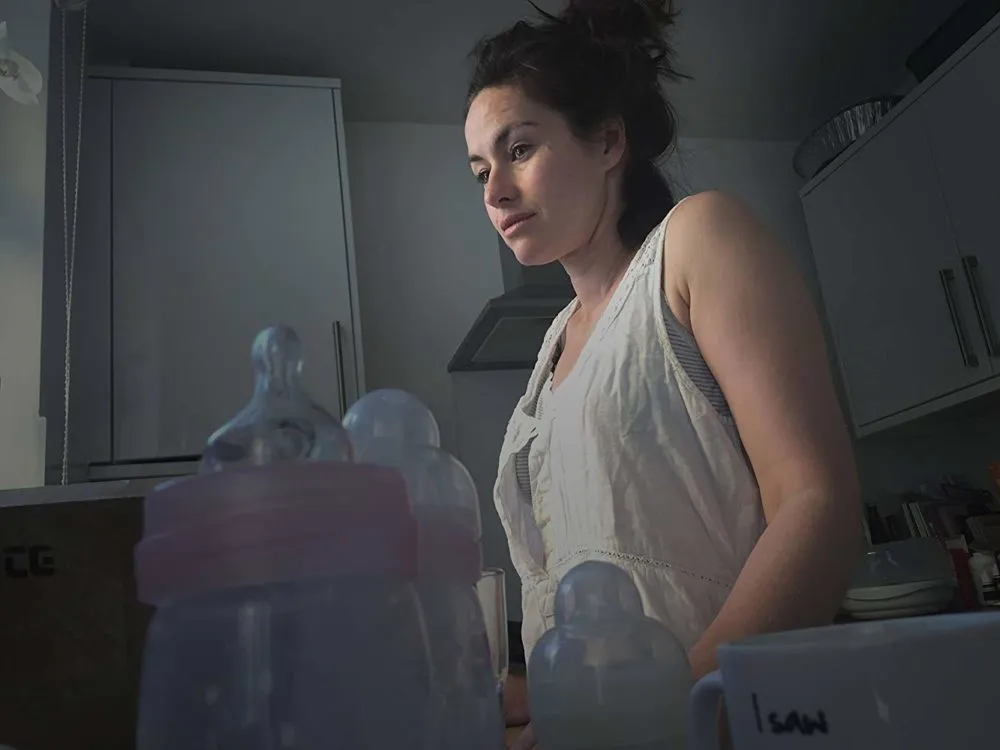 A woman stands in a dimly lit kitchen with baby bottles and a labeled container in front of her.