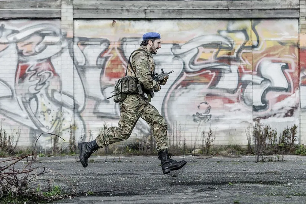 A soldier in camouflage uniform runs with a gun in front of a graffiti-covered wall.