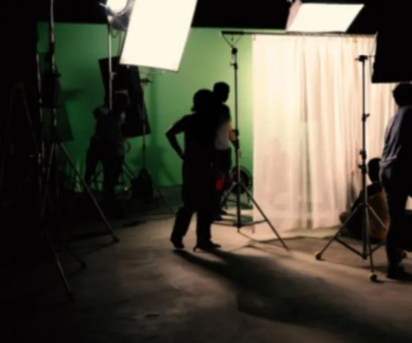 Film crew arranging lighting and equipment on a green screen set with a white curtain in the background.