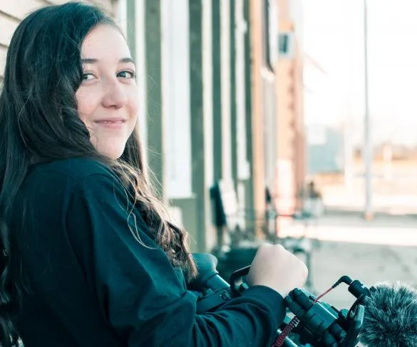 Person with long hair smiling at the camera, holding video equipment outdoors near a building.