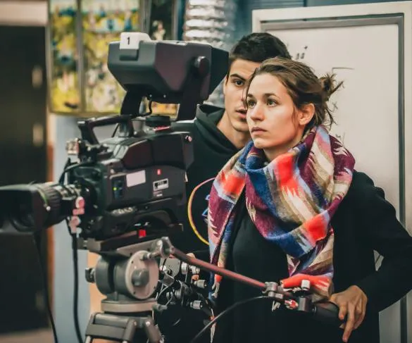 A woman wearing a colorful scarf operates a professional video camera on a tripod, with a man standing closely behind her.