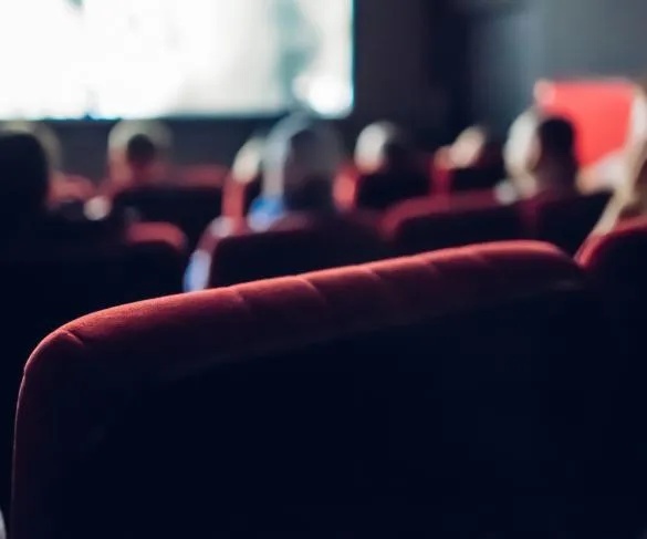 People sitting on red seats in a dimly lit cinema, facing a blurry screen in the background.