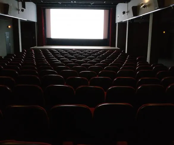 An empty movie theater with rows of red and black seats facing a large illuminated white screen.