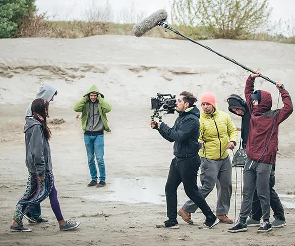 Film crew operating camera and boom mic while recording two actors outdoors in a sandy area; another person stands in the background.