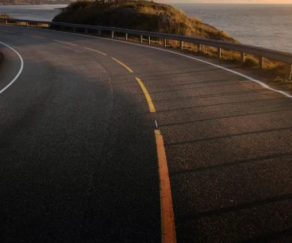 A curved coastal road with two yellow lines, metal guardrails, and a view of the ocean under a setting sun.