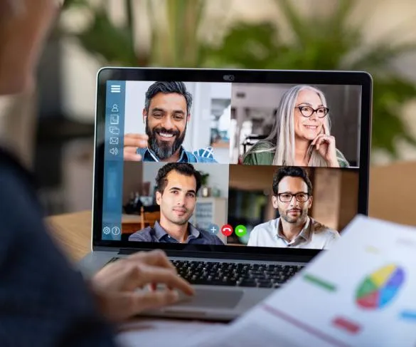 Four people are engaged in a video conference call on a laptop screen, with a paper showing a pie chart in the foreground.