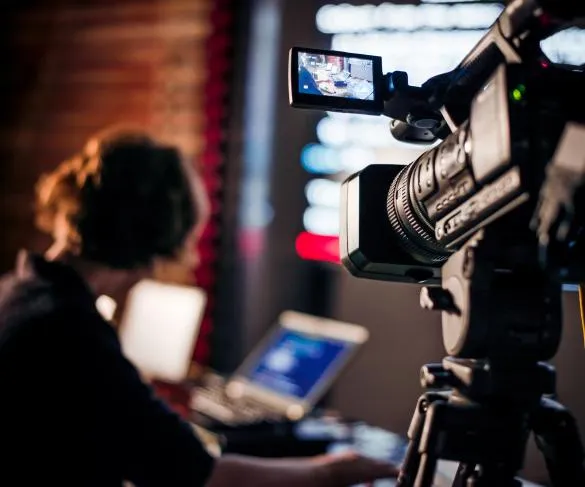 A person sits at a desk operating a video camera with a screen displaying a video feed.