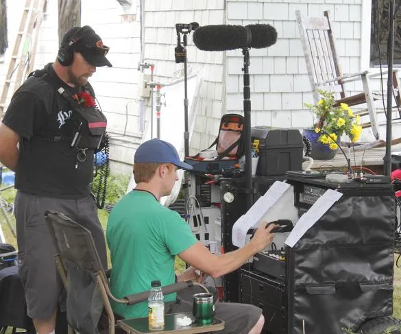 Two men operate sound equipment outdoors, with one adjusting settings and the other wearing headphones. Equipment and notes are visible.