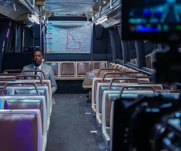 A man in a suit sits alone on a mostly empty bus with a city transit map displayed in the background and a camera visible in the foreground.