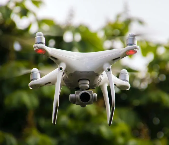 A white quadcopter drone with a camera hovers outdoors against a blurry green foliage background.