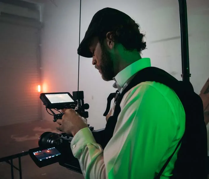 Person wearing a hat and waistcoat operates a camera on a film set with green lighting.