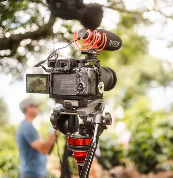 Close-up of a video camera on a tripod with a Rode microphone attached, set up outdoors. Blurred person and greenery are in the background.