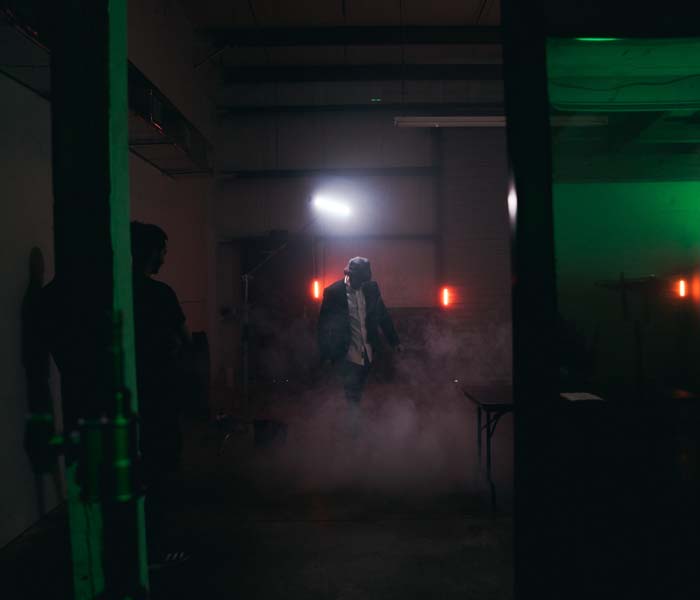 Person standing in a dimly lit room filled with smoke, illuminated by red and green lights, with filming equipment in the background.