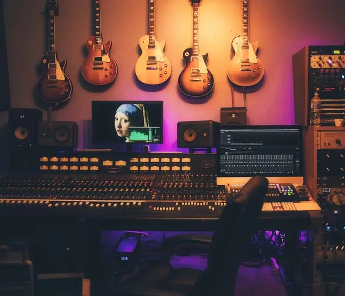 A music studio with a control board, computer, and a screen displaying artwork. Guitars hang on the wall in the background.