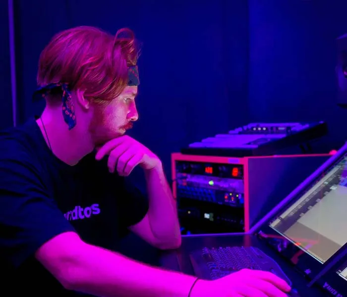 Person with long hair and headband working in a dimly lit room, looking at a computer screen with audio equipment in the background.