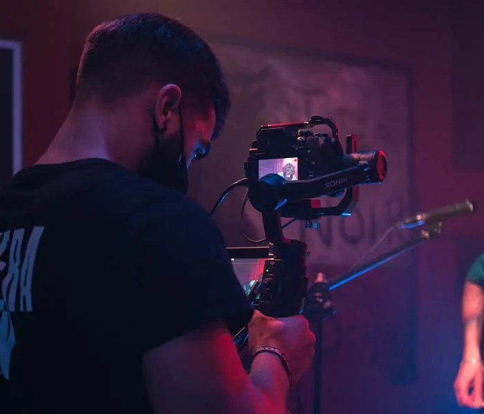 Person wearing a mask is filming with a camera on a stabilizer in a dimly lit room, with a microphone stand visible in the background.