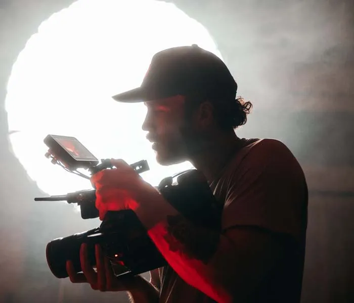 A man wearing a cap operates a camera with a bright light source behind him, creating a silhouette effect.