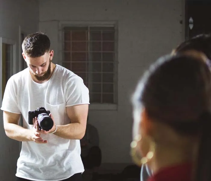 A man holding a camera points it towards a person in the foreground with blurred focus.