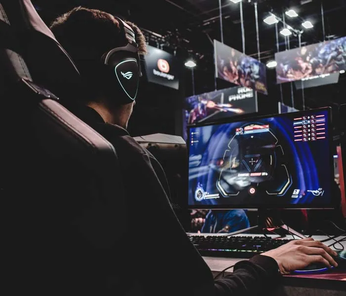 Person wearing headphones and sitting in front of a computer monitor while playing a video game in a dimly lit gaming area.