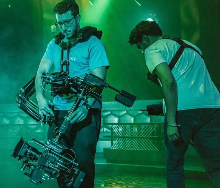 Two camera operators work with stabilization rigs in a dimly lit room with green ambient lighting.