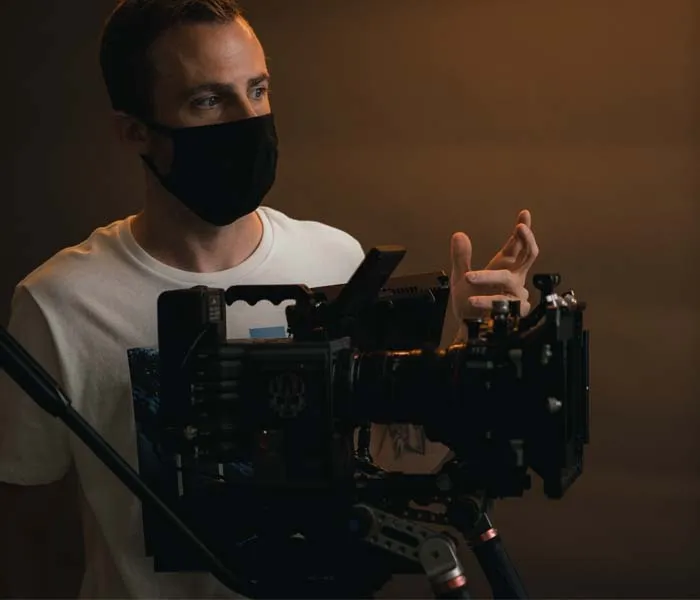 Person wearing a black mask operates a professional camera setup on a tripod in a dimly lit environment.