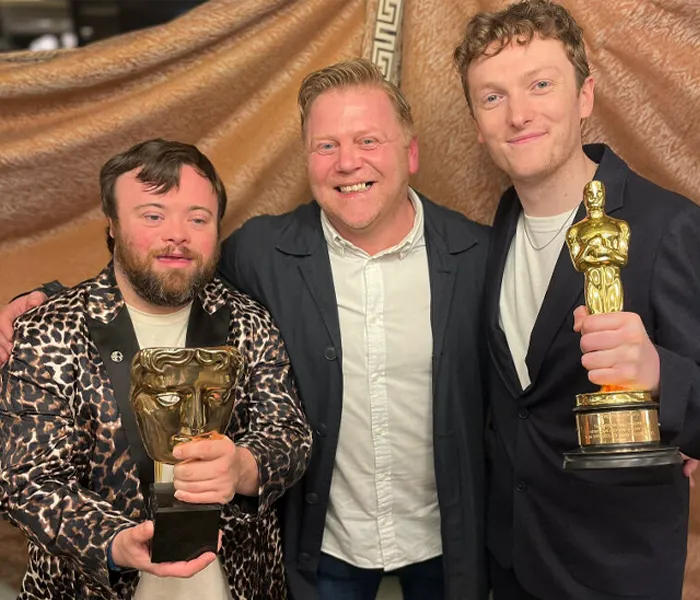 Three men are standing together, smiling, with two of them holding awards. The man on the left holds a BAFTA trophy, and the man on the right an Oscar.