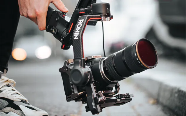 A person holds a camera equipped with a stabilizer while crouching on a sidewalk, close-up shot focused on the camera.