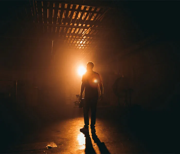 A person stands in a dimly lit room with a bright light shining behind them, casting a long shadow on the floor.