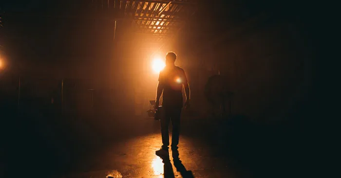 Silhouetted person walking in a dimly lit space with bright orange lights in the background.