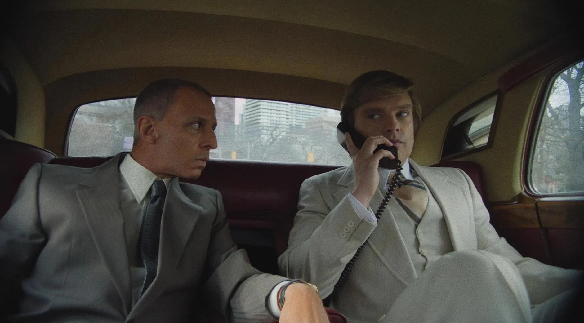 Two men in suits sit in the backseat of a car. One is talking on a car phone while the other looks at him. City buildings are visible outside.