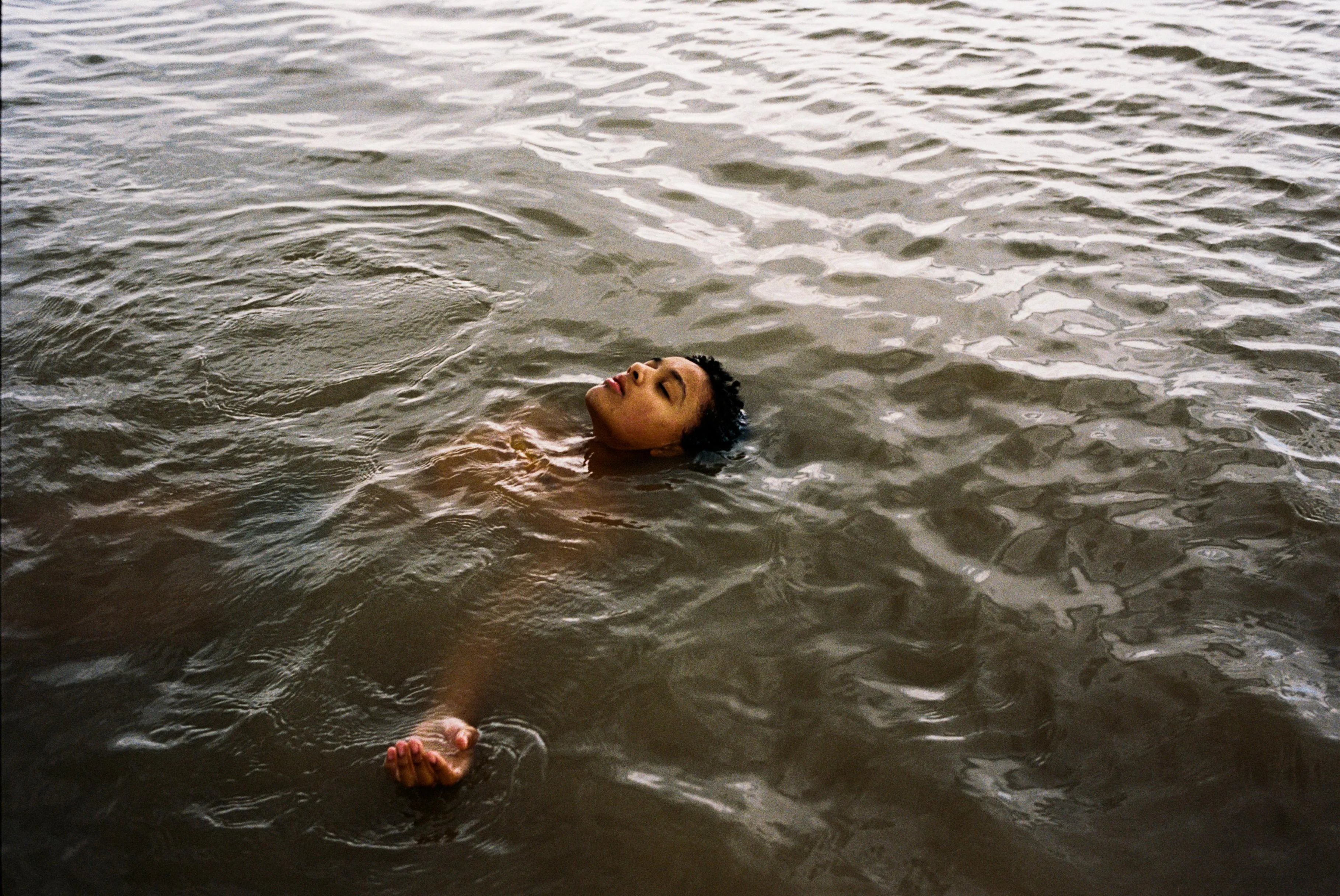 A person floating on their back in calm, shallow water, with eyes closed and one arm extended to the side.