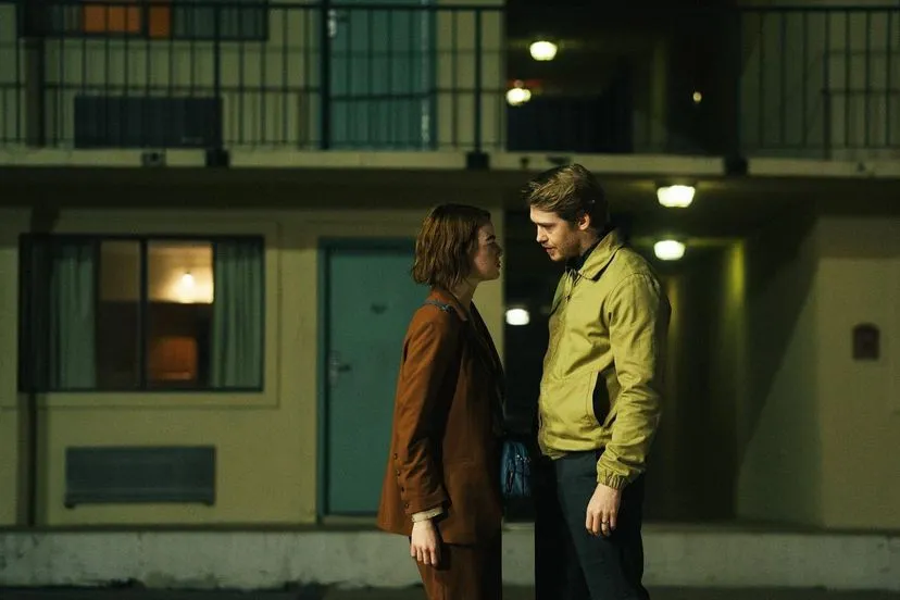 Two people are standing outside an apartment complex at night, conversing closely under dim lights.