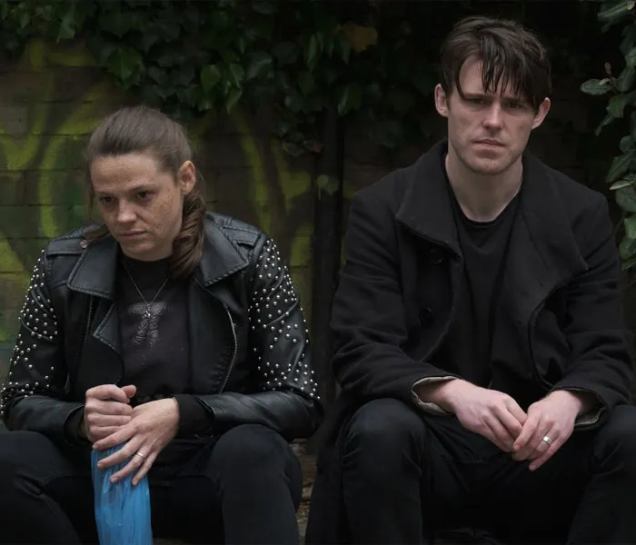 Two people sit side by side against a graffiti-covered wall, both wearing dark clothing and appearing somber.