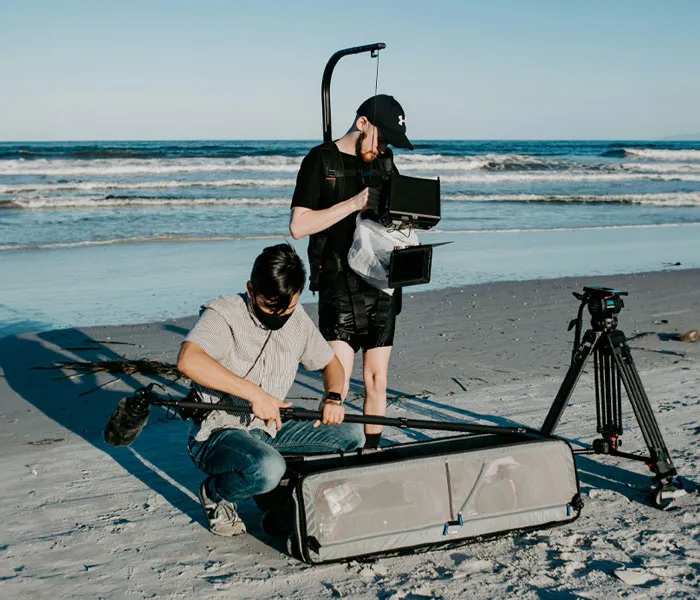 Two people setting up film equipment on a beach; one person handles a boom mic, and the other adjusts a camera monitor.