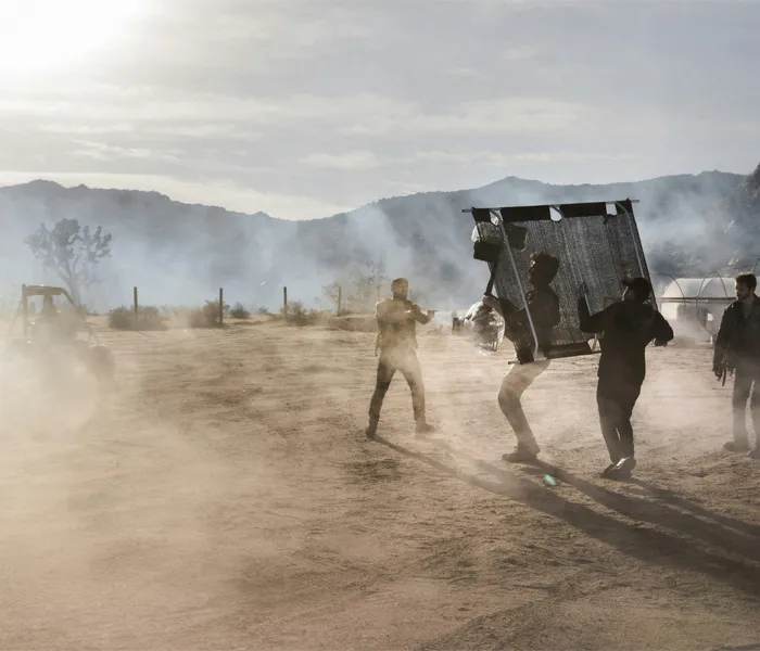 People in a dusty outdoor setting using a large fabric screen for shade or wind protection on a sunny day with mountains in the background.