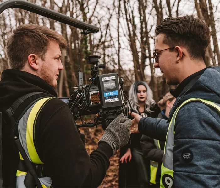 Two crew members operate a camera on a film set in a forest. An actor in costume stands in the background.