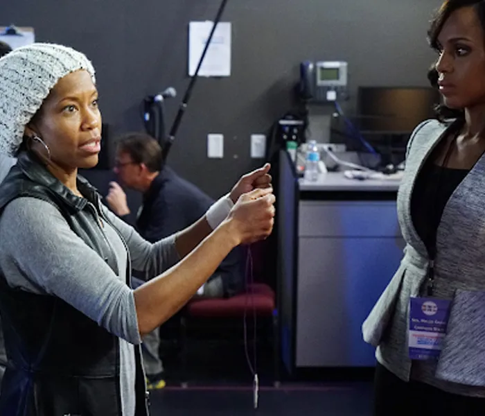 Two women engage in a conversation backstage at an event. One is holding a cord, while the other listens intently.