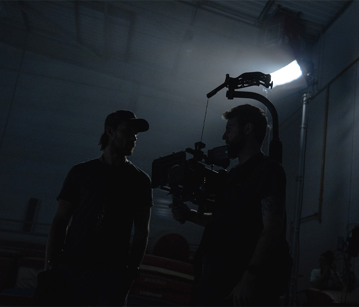 Two men stand in a dimly lit room, illuminated by a single light source, creating a dramatic atmosphere.