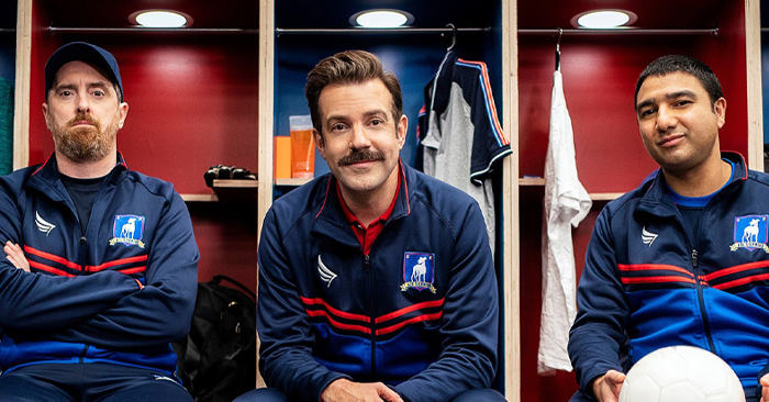 Three men in a locker room, casually sitting together, with a soccer ball positioned nearby on the ground.