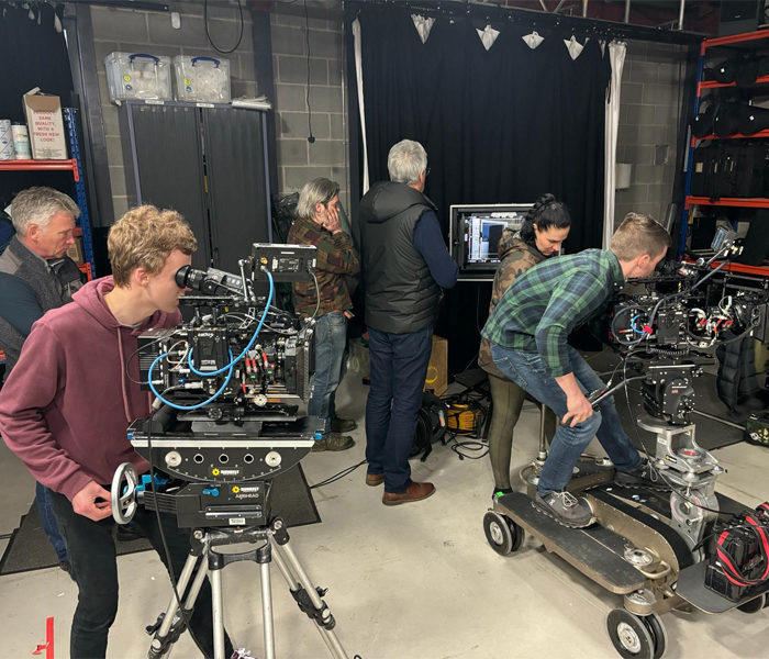 Group of people operating film cameras and equipment in a studio space with grey walls and various supplies on shelves.