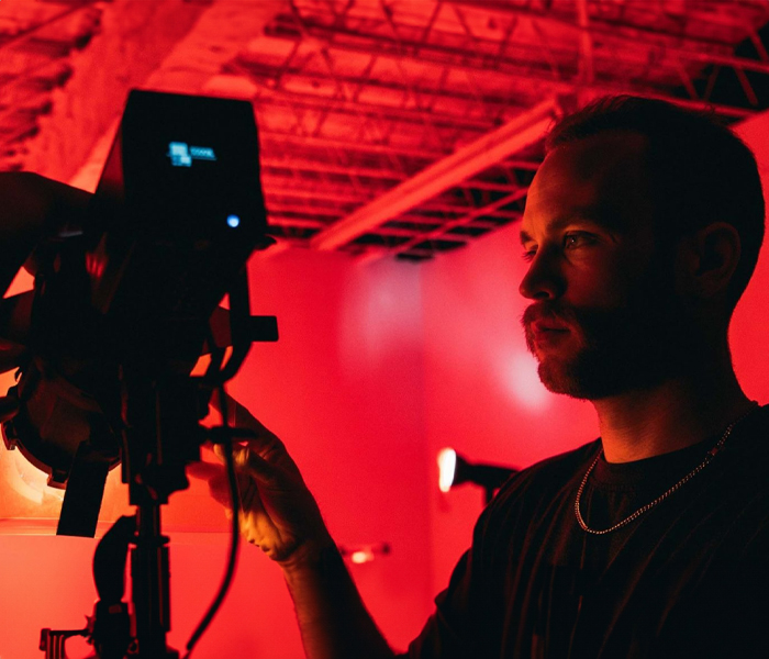 Person in a dark room, illuminated by red light, adjusting settings on a camera mounted on a tripod.