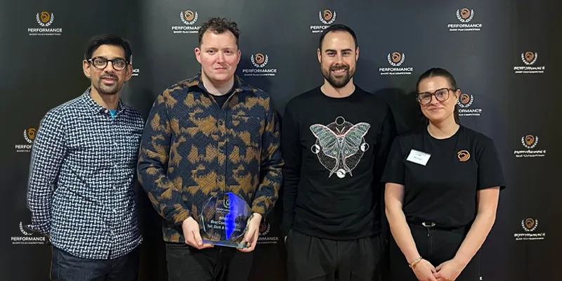 Four people standing together; one is holding an award. They are in front of a backdrop with "Performance" logos.