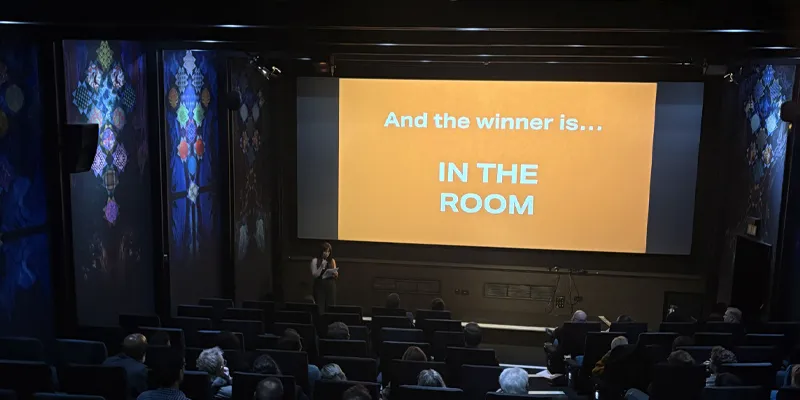 A person stands in front of a screen displaying "And the winner is... IN THE ROOM" in a dimly lit theater with an audience seated.